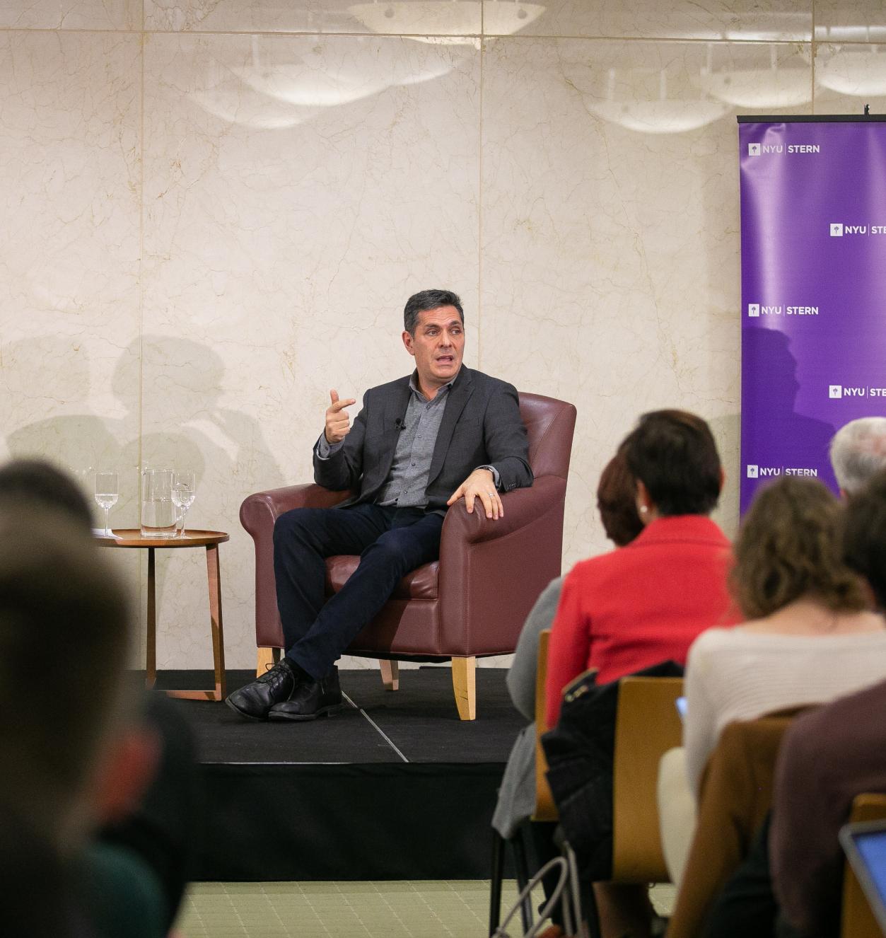A woman and a man seated in armchairs on a raised platform in front of an audience. NYU banner on the right-hand side