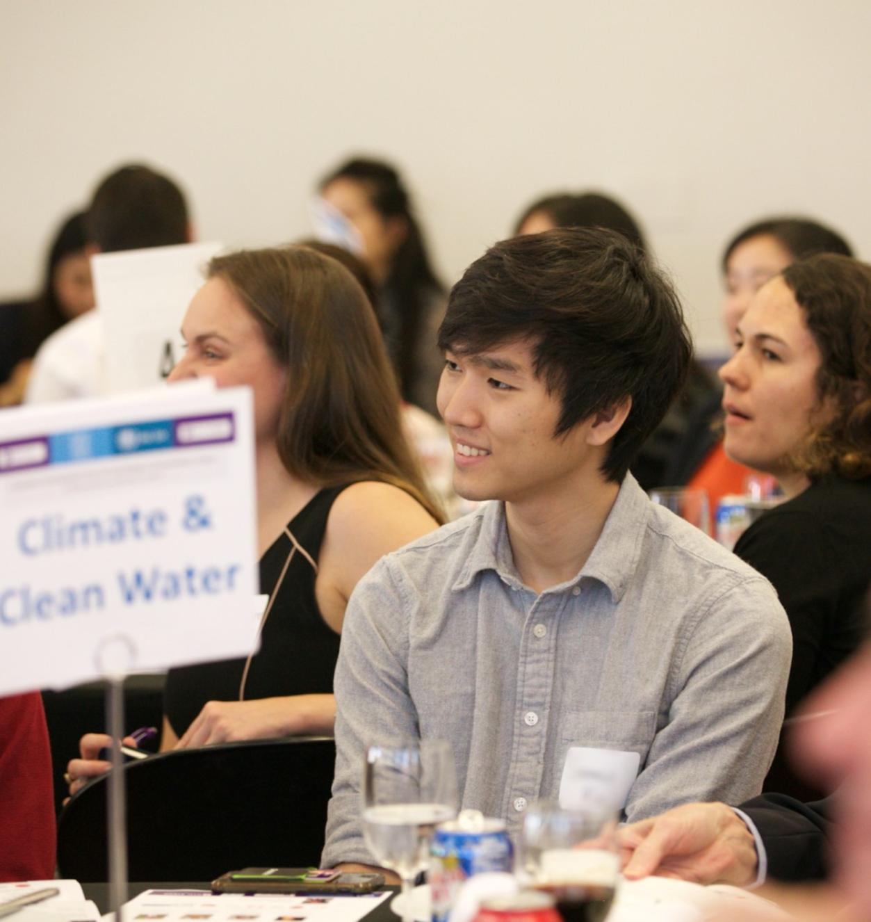 Young man in focus sitting at a table with a sign reading Climate & Clean Water. 