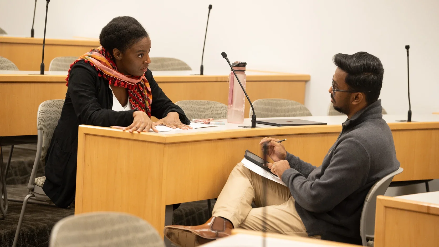 A feminine-presenting person with dark skin wearing a black pant suit and red rivulet of a scarf sits above a masculine-presenting person with dark skin wearing a gray cardigan and khakis. She gives him a serious look; it looks like she's laying down the law with her confident posture and serious gaze.