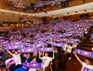 A photo of a big crowd of NYU Stern Undergraduate Students sitting in an auditorium and holding signs that say "Stern."