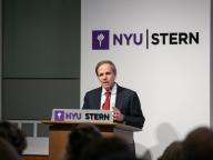 A photo of Michael Posner speaking at a podium that says NYU Stern. He is wearing a suit with a red tie.