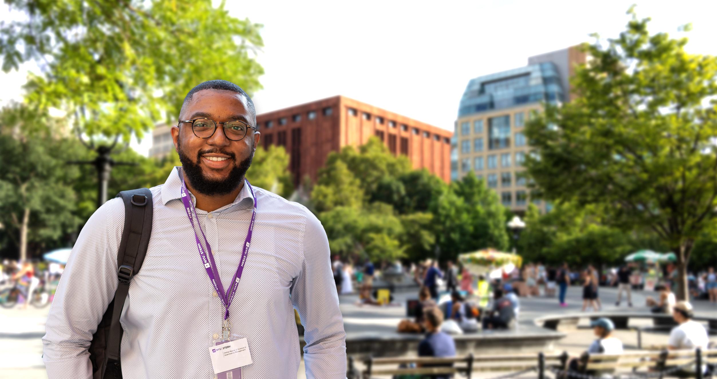 Male student with backpack, light color shirt and a New York University ID hanging from his neck in front of blurred background image of Stern School of business seen from the fountain at Washington Square Park