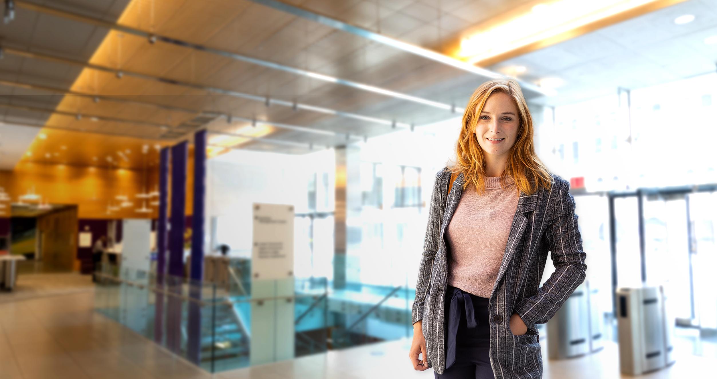 Female student with red hair, grey coat and pink blouse in front of a blurred background of Stern's lobby floor