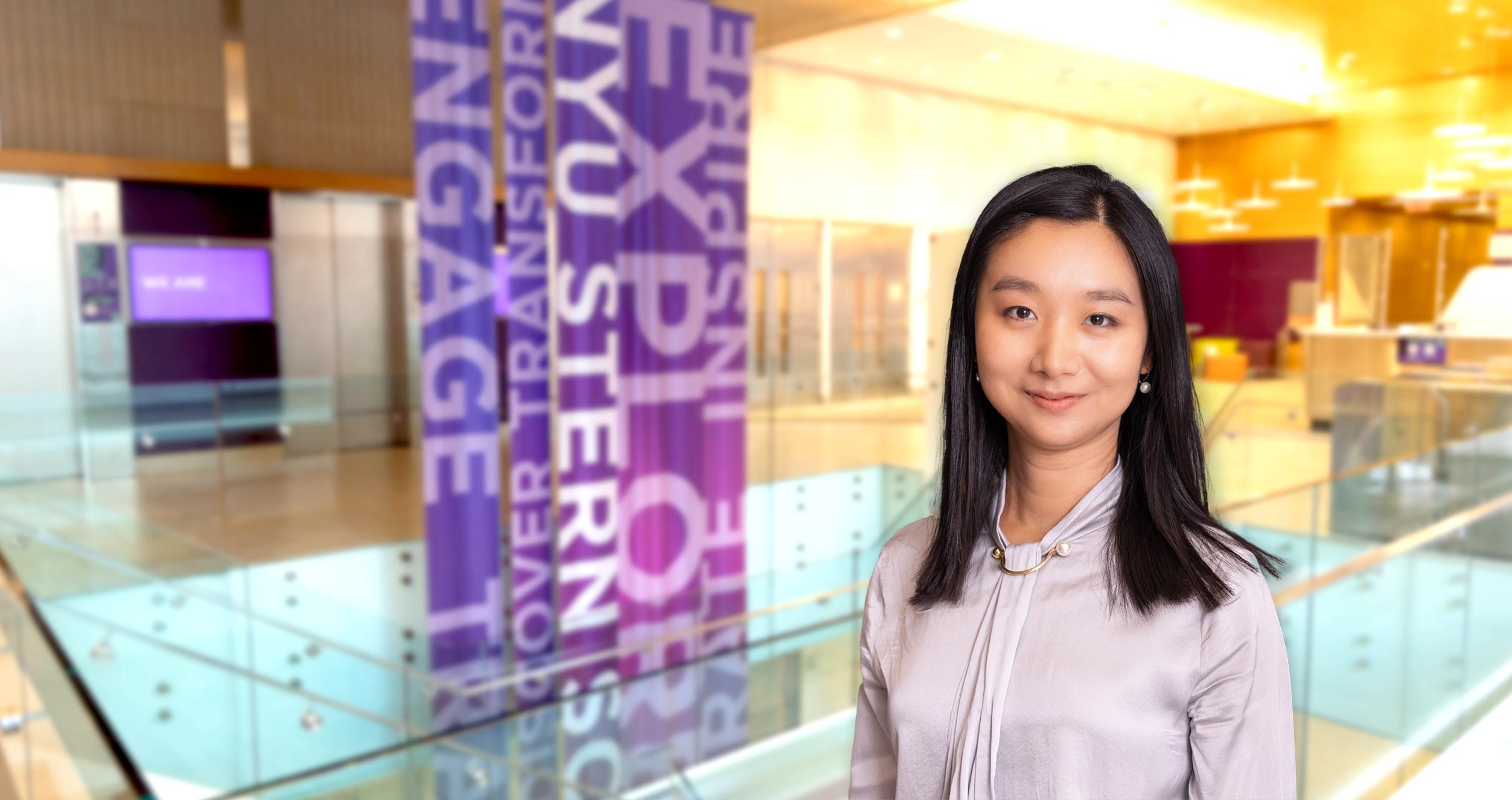 Female student with dark color hair and light color shirt in front of a blurred background of Stern's lobby floor