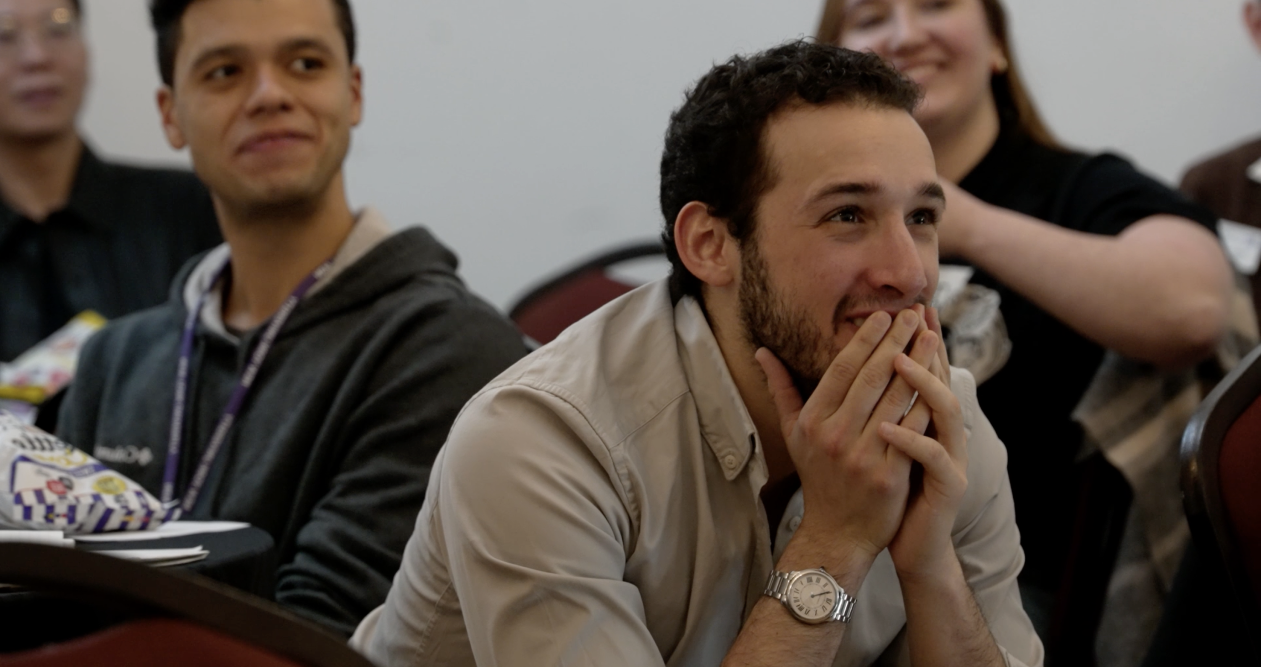 A masculine presenting person with short curly hair sits in a crowd of people, hands covering his smile in excitement and attentive listening. 