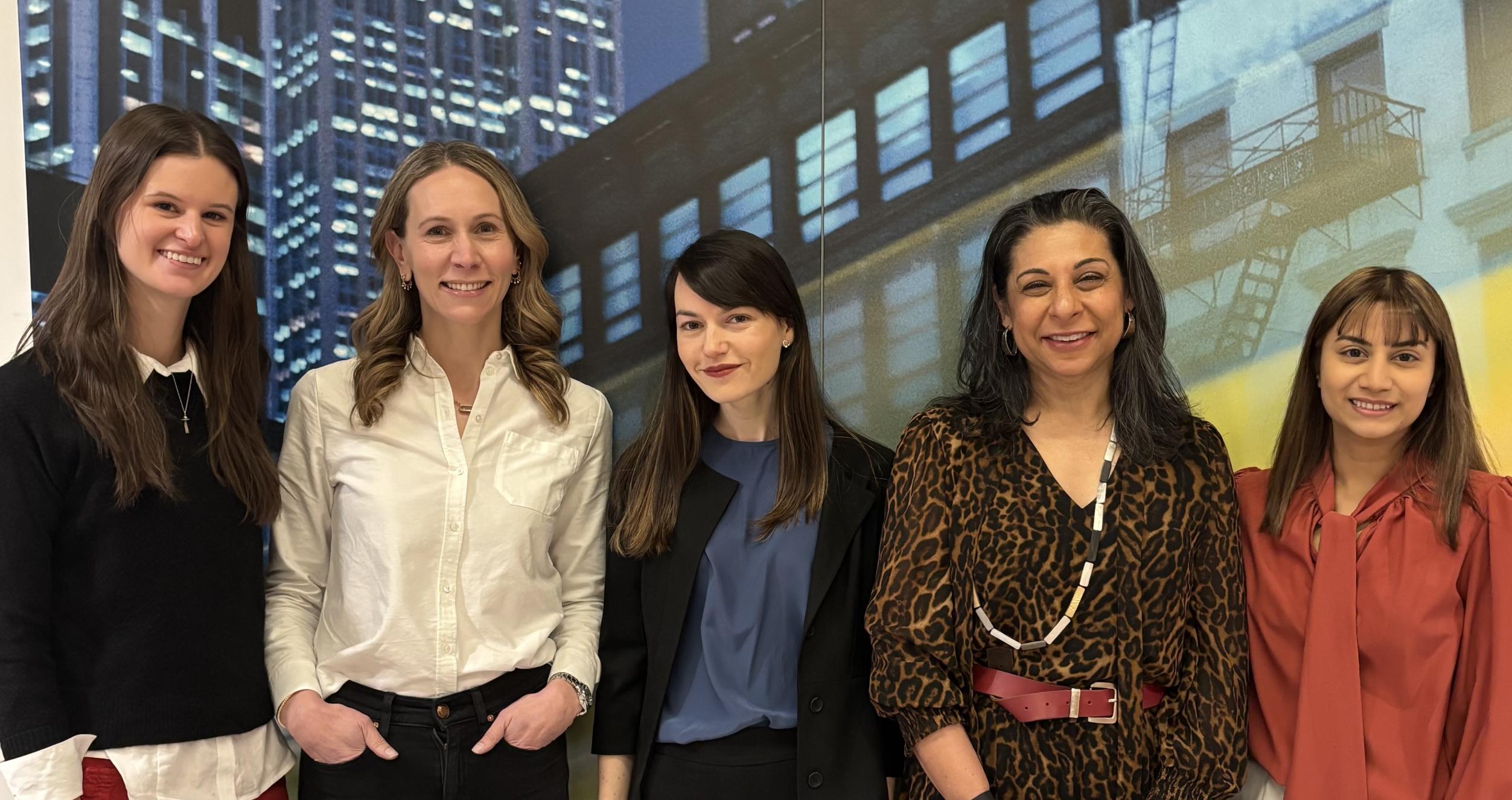 A group of five feminine presenting people line up against a background of city-scape wallpaper.