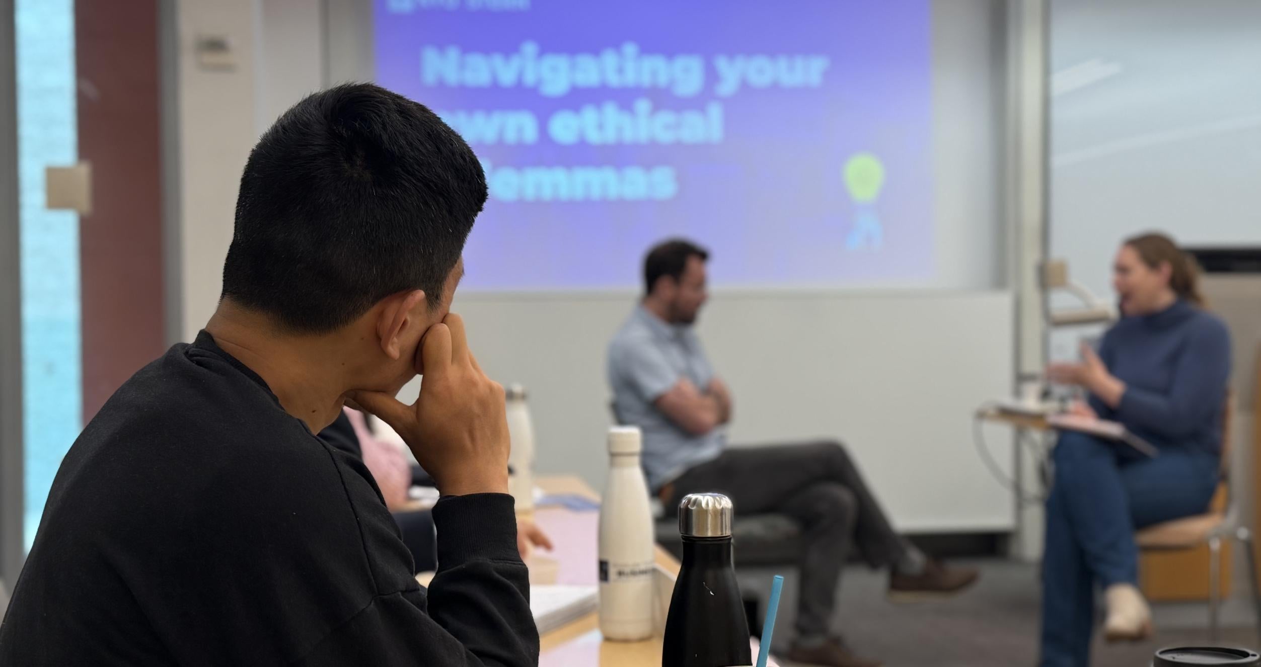 A masculine-presenting student with dark skin and hair looks thoughtfully at a pair of actors in discussion at the front of the class. A screen reads, "Navigating your own ethical dilemmas" above the actors' heads.
