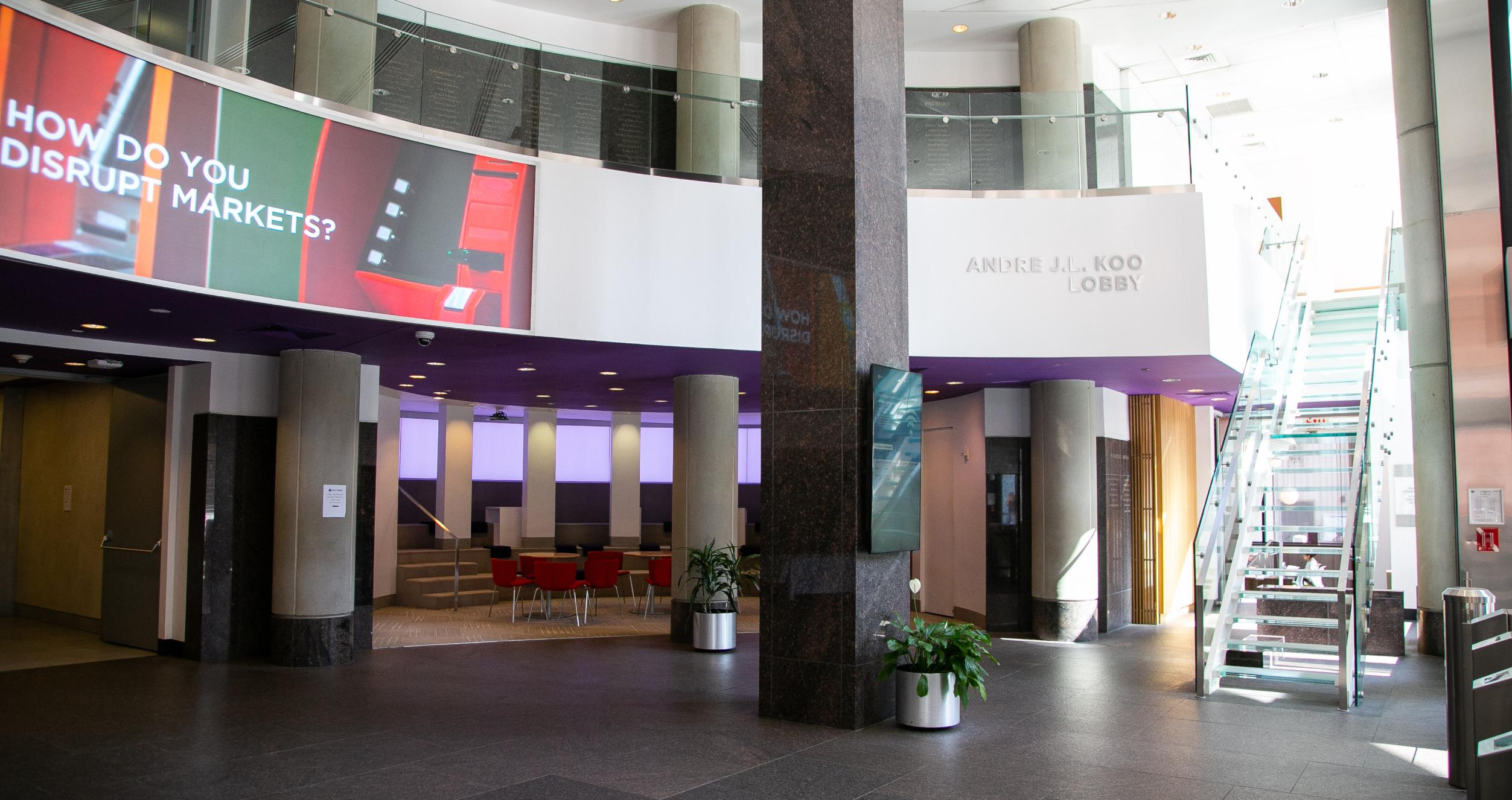 Ground floor view of empty lobby, clear stairs going up to a second level, column in the center and large screen in upper leff