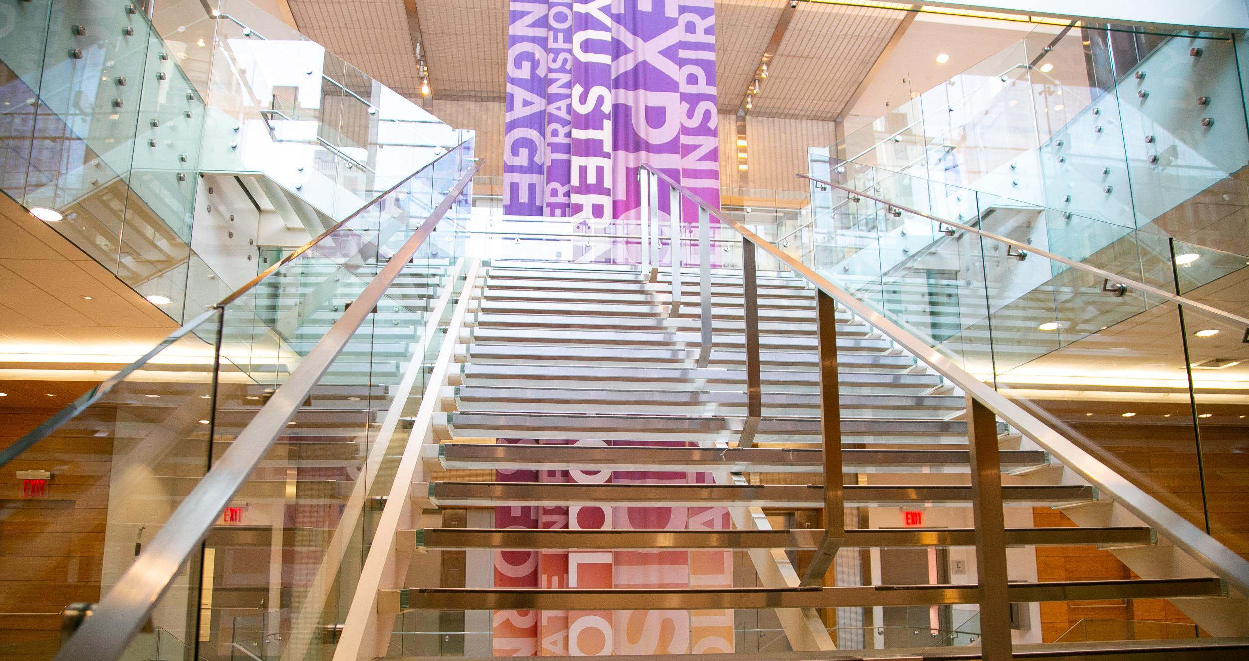 View from the bottom of a modern-looking glass stairway with a multi-colored banner behind it, located in Tisch Hall. 
