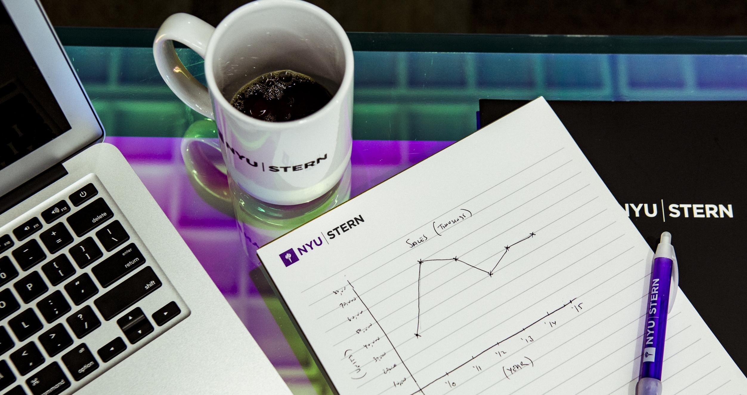 A laptop, notebook with a pen and a mug on a desk