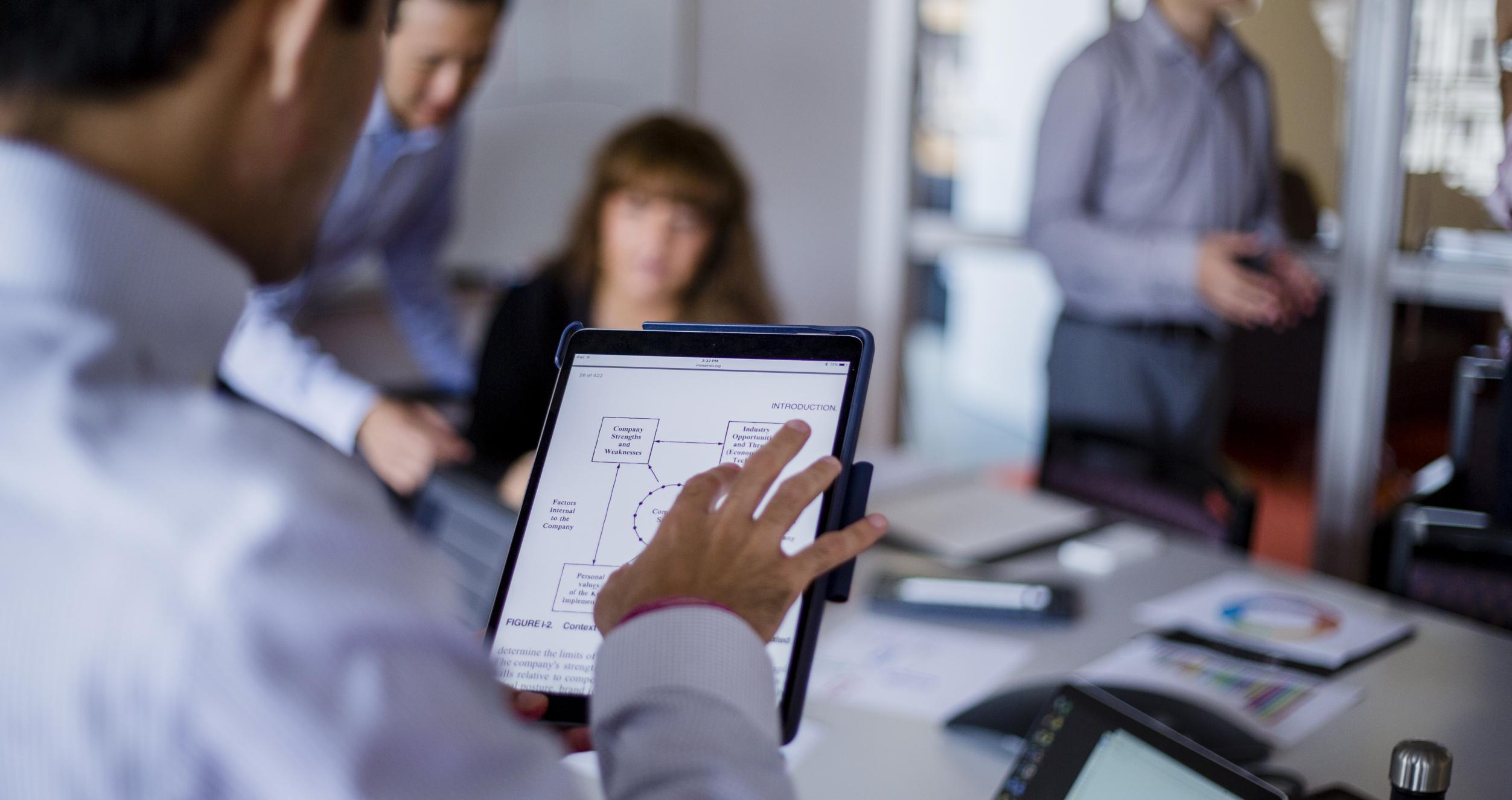 Image of a small meeting, we see over the shoulder of a man holding a tablet, and in the background are 3 other employees. 