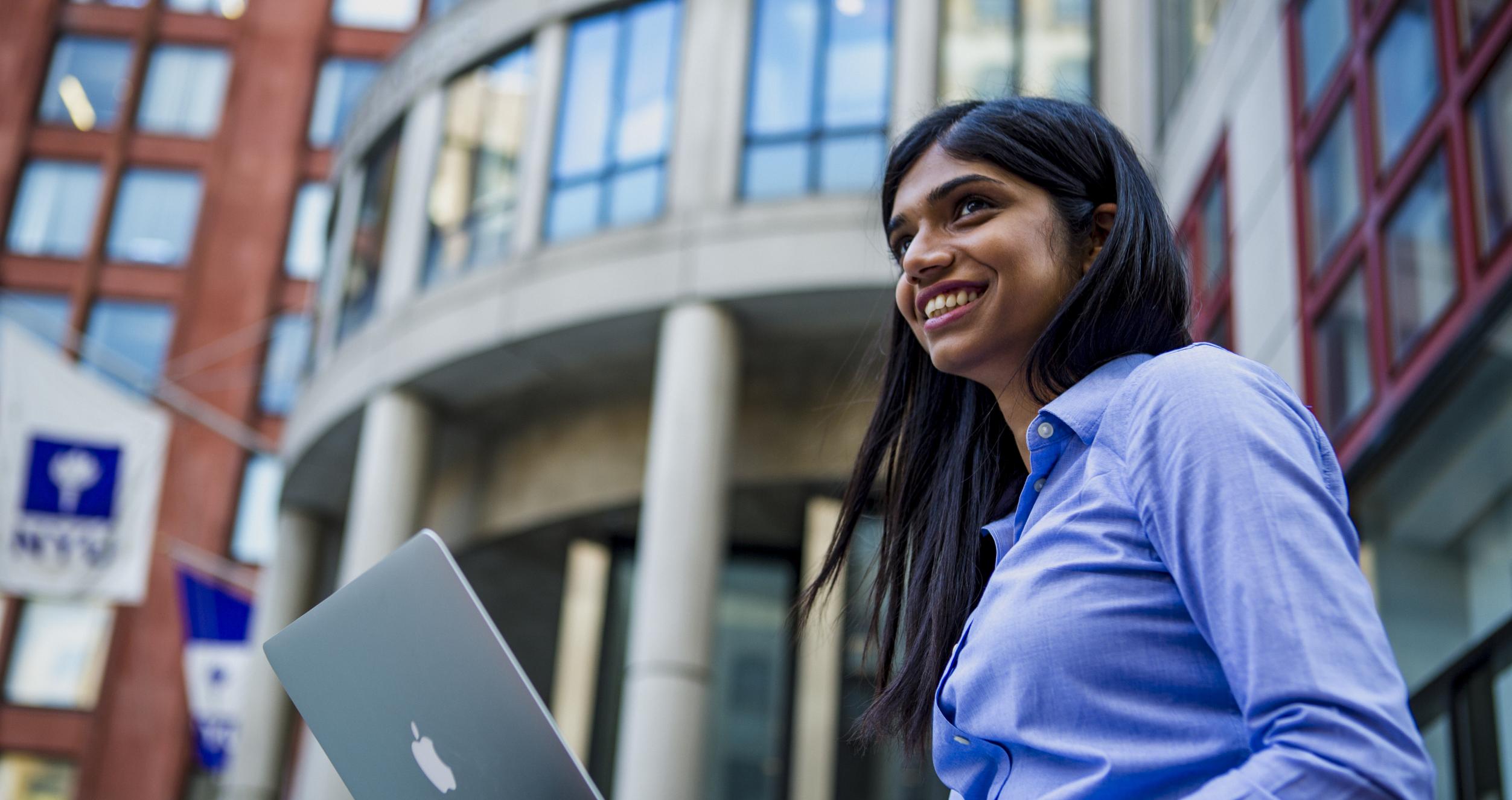 Smiling young woman sitting in front of Stern building with a laptop on her lap