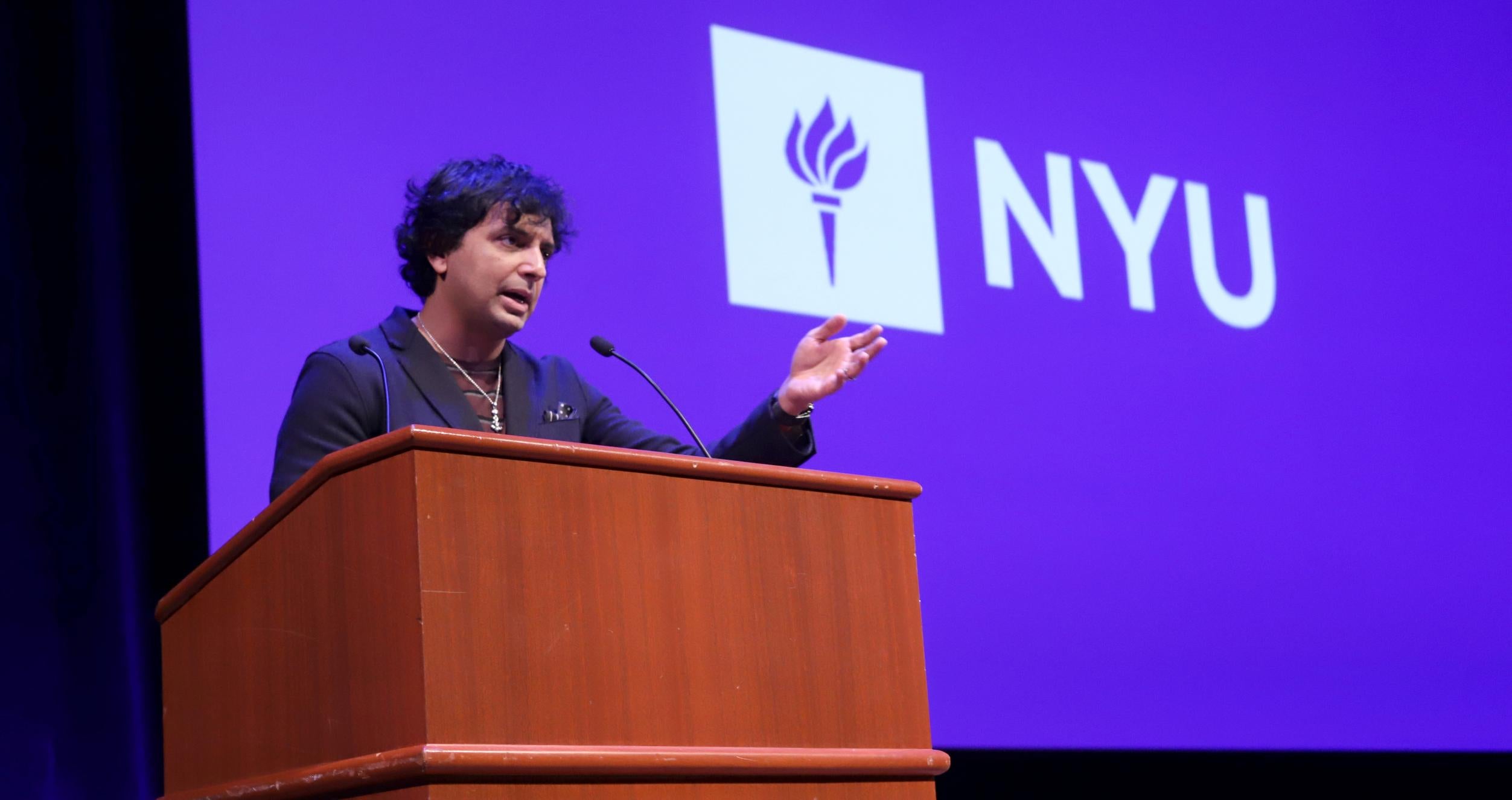 Filmmaker M. Night Shyamalan speaking at a podium in front of NYU logo background