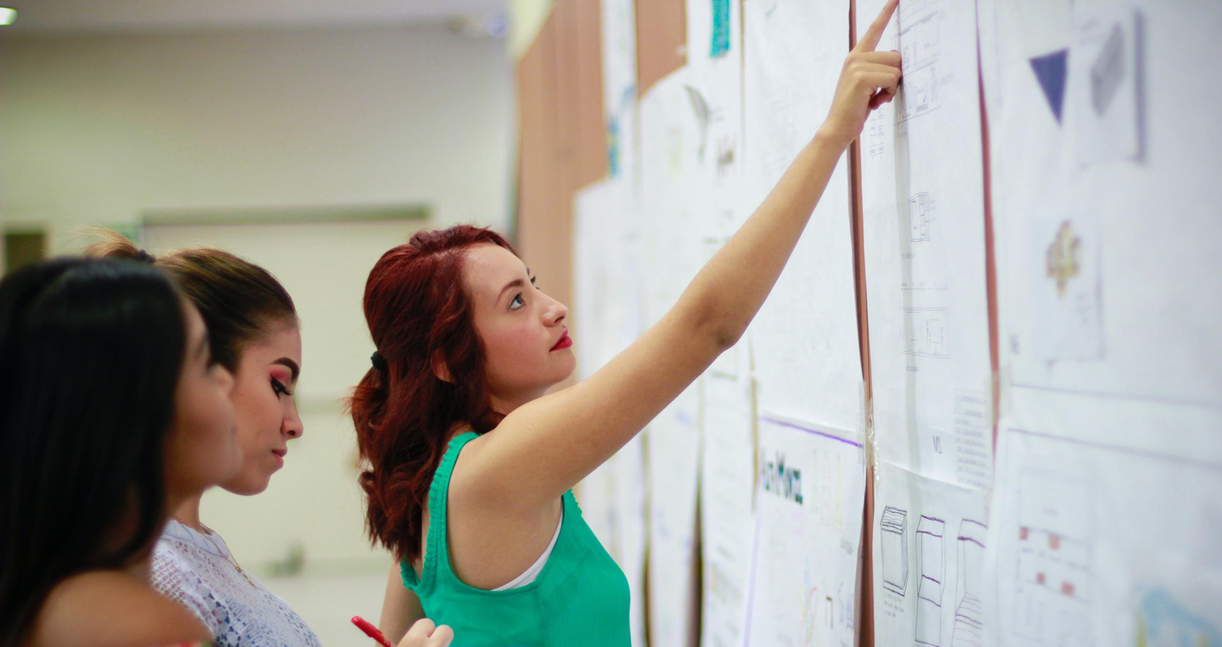 Three young women in front of a bulletin board filled with papers. One woman is pointing at something on the board.