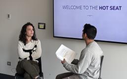 A feminine-presenting person with fair skin and curly, shoulder-length hair sits with her arms crossed across from a masculine-presenting person with medium-tan skin and dark hair, as he olds a piece of paper in his hands and gestures while talking. A screen behind these two reads, "Welcome to the hot seat."