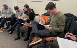 Five students sit in a row writing out responses on pieces of paper that sit on their laps.