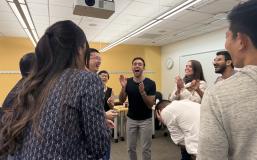 A group of seven students stands in a circle clapping and laughing after a game of rock paper scissors.