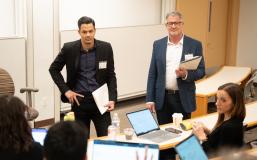 Two masculine presenting people with lighter skin stand in blazers as they hold a pile of papers. One stands with his right hand on his hip. They listen to a small group of seated people in a lecture-style classroom.  