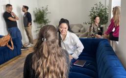 Three pairs of students sit engaged in conversation in a room featuring two large blue couches, two comfy linen chairs, and two large house plants. On the far left of the photo, two masculine-presenting students lean against the doorframe as they talk. In the center forefront, two feminine-presenting people sit on a blue couch, one with her back to the camera and the other, smiling as she looks at her partner, facing the camera. On the far right, two feminine-presenting converse in the windowsill.