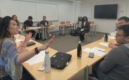 A group of twelve students addresses a feminine-presenting actor with dark hair and skin seated in the middle of the room. 