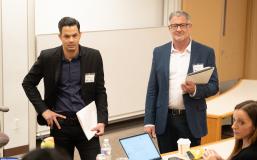 Two masculine presenting people with lighter skin stand in blazers as they hold a pile of papers. One stands with his right hand on his hip. They listen to a small group of seated people in a lecture-style classroom.  