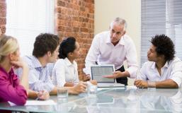 four people sitting at a glass table looking at a fifth person who is explaining something from a laptop