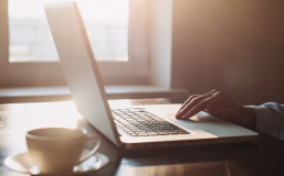 hand on touchpad of a laptop, next to a mug, in a room with sunshine beaming in