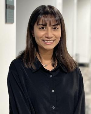 Aasiya Van A feminine-presenting person with medium-dark skin tone, and short, straight black hair, and bangs smiles into the camera. She is wearing a black silk collared shirt and standing in the middle of a long empty hallway.