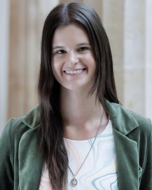 Ellie Houlihan A feminine-presenting person with long brown hair parted down the middle smiles into the camera. They are wearing a velvet green blazer and white crewneck shirt with thin wavy lines of various colors dancing along the bodice. They are wearing a long silver necklace, and they are standing in front of a row of large Greek columns.