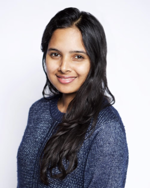 A feminine presenting person with dark hair and medium tan skin smiles into the camera. She wars a thin cable-knit sweater and her hair falls in curls long past her shoulder.