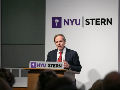 A photo of Michael Posner speaking at a podium that says NYU Stern. He is wearing a suit with a red tie. 