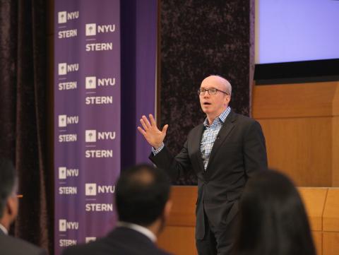 A man speaking to an audience in front of NYU banners