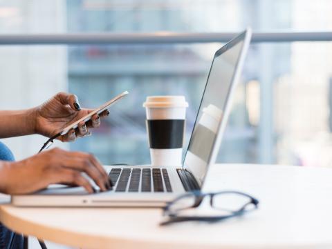 Person's hands on a phone with a laptop, cup of coffee, and pair of glasses in the frame