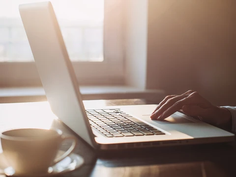 hand on touchpad of a laptop, next to a mug, in a room with sunshine beaming in