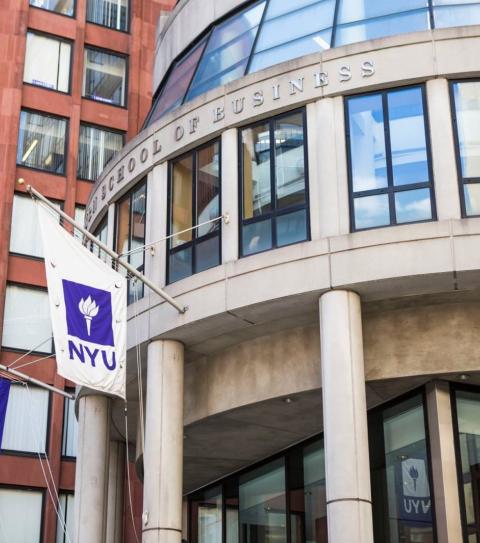 A photo of the outside of NYU Stern. It is a rounded concrete building with NYU flags hanging outside.