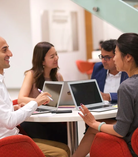 Four students seated around a round table, each with laptops in front of them, engaged in conversation.