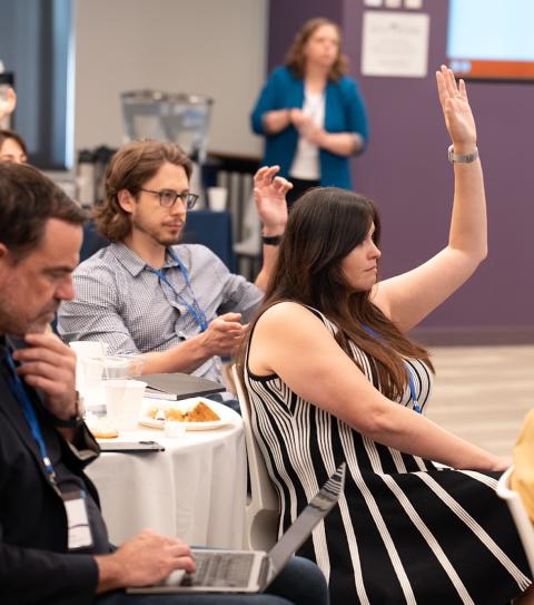 several people raise hands at a table watching a speaker