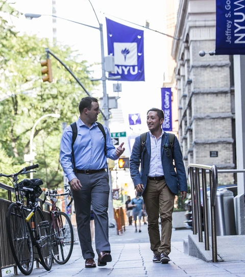 Two men walking towards the camera in conversation, NYU banners behind them