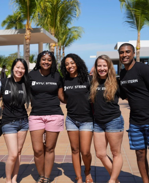A group of NYU Stern students stands by a pool in black NYU Stern tshirts smiling.