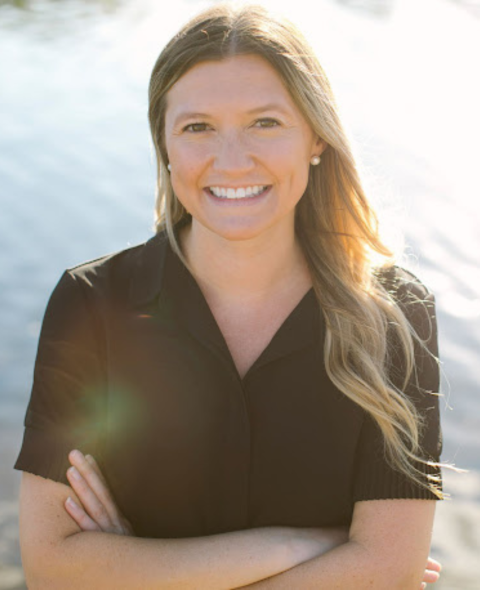 Anna has blonde wavy hair. She stands in a black shirt smiling at the camera in front of a body of water.