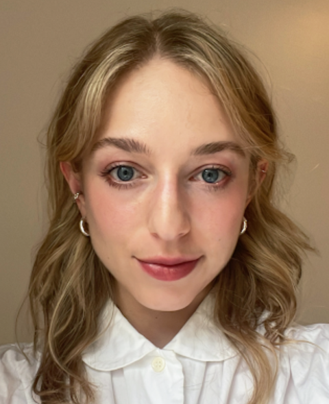 Rose stares directly at the camera. She has wavy blonde hair and wears a white button down against a tan background.