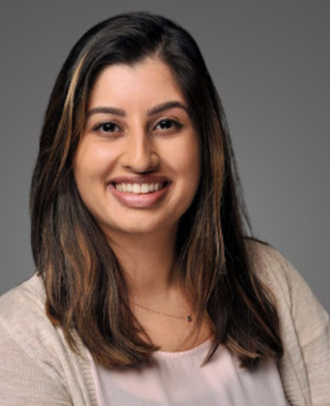 Zoish has brown hair and wears a tan shirt. She smiles at the camera against a gray background.