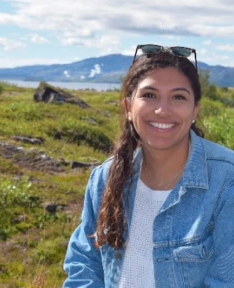 Cecilia sits in front of a mountain with a jean jacket. She has long brown hair and smiles at the camera.