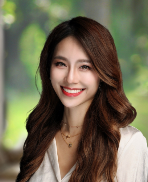 Christy Kim's headshot. She has wavy long brown hair and wears a white shirt. She smiles at the camera against a green background.
