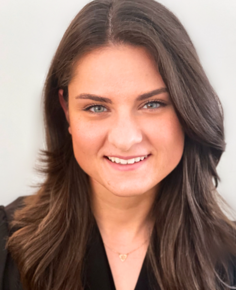 Carina has wavy brown hair and smiles at the camera in a black top against a white background.