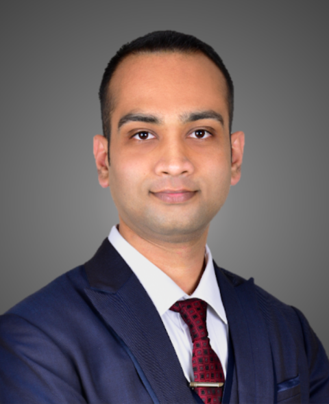 Shiriam's headshot. He stares at the camera and wears a blue jacket and red tie against a gray background.