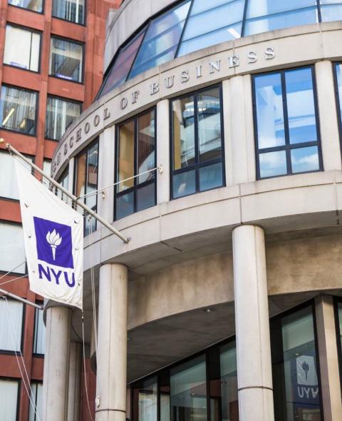 A photo of the outside of NYU Stern. It is a rounded concrete building with NYU flags hanging outside.