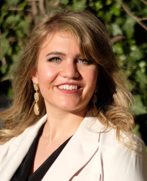 Isabella stands outside smiling at the camera. She has wavy blonde hair and wears a black blouse and tan blazer.