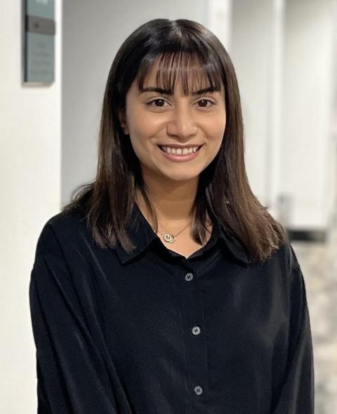 Aasiya Van A feminine-presenting person with medium-dark skin tone, and short, straight black hair, and bangs smiles into the camera. She is wearing a black silk collared shirt and standing in the middle of a long empty hallway.