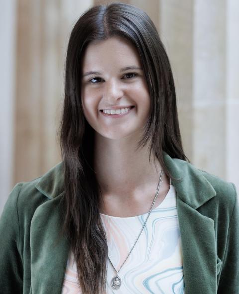 Ellie Houlihan A feminine-presenting person with long brown hair parted down the middle smiles into the camera. They are wearing a velvet green blazer and white crewneck shirt with thin wavy lines of various colors dancing along the bodice. They are wearing a long silver necklace, and they are standing in front of a row of large Greek columns.
